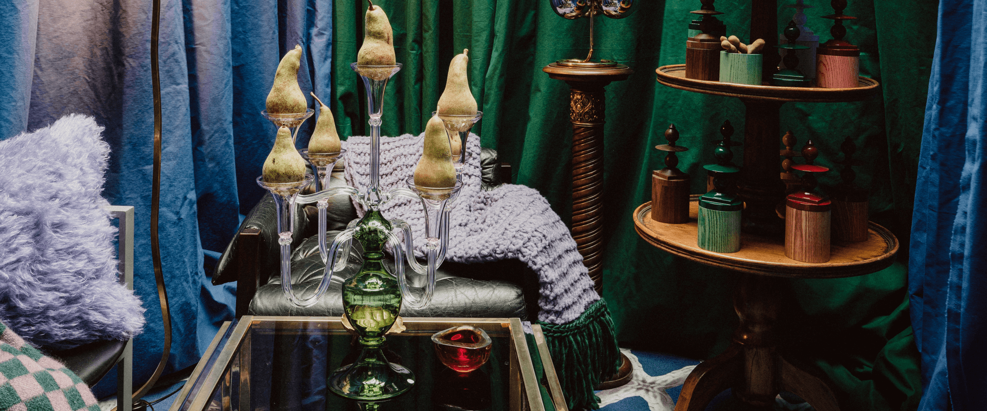 Hand-blown glass candelabra with pear-shaped ornaments displayed on a glass table in a richly layered interior — part of the Gabriela Seres Home Accents collection, showcasing sculptural glass pieces in warm light and textile textures.