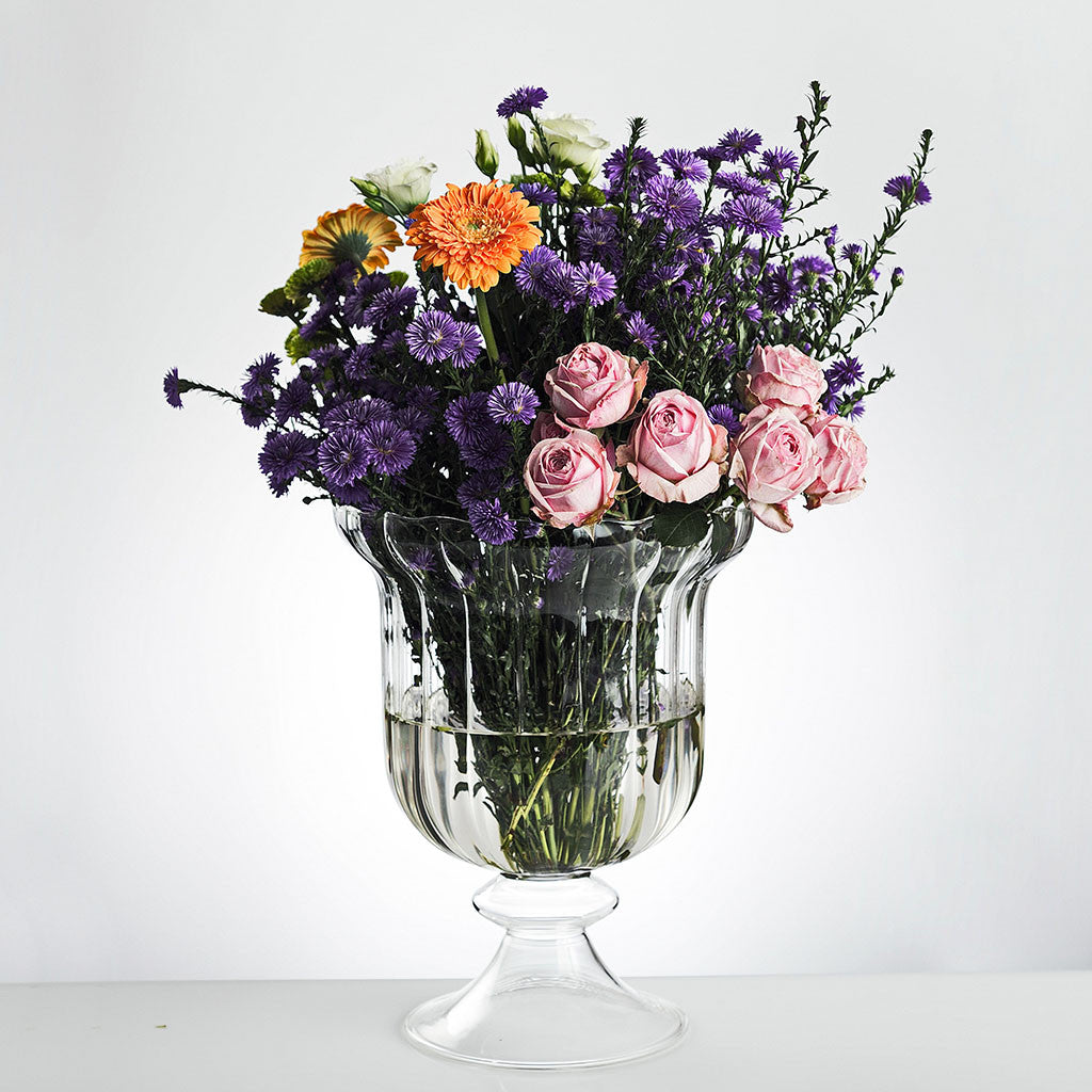 A wide, clear glass vase with a bouquet of multicolored flowers arranged in it, set against a white background.
