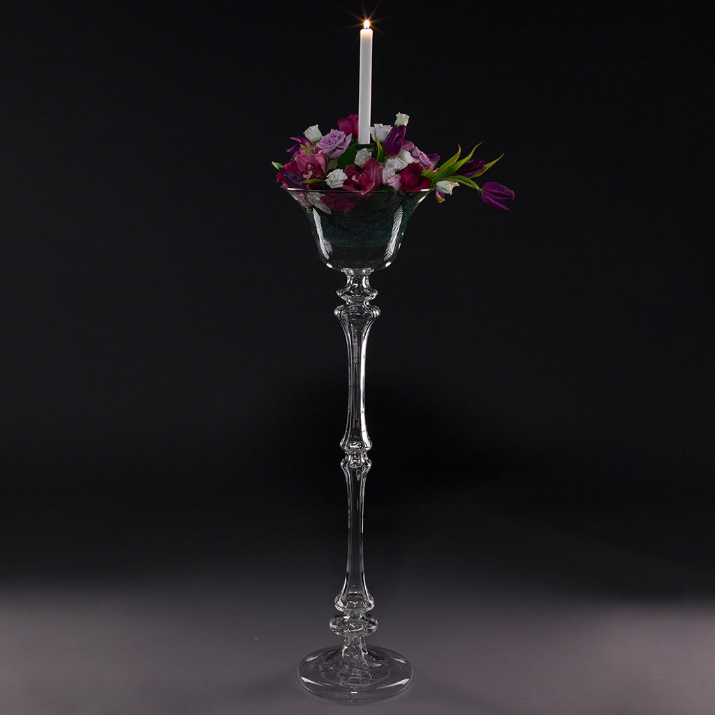 A clear glass flower stand with a floral arrangement in it, featuring a lit candle at the top.